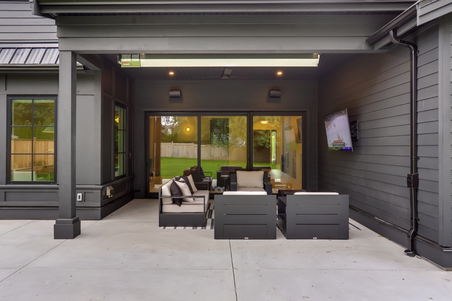 Covered outdoor living room with dark exterior siding, skylight, wide sliding glass doors, mounted TV, and lounge seating on a concrete patio.