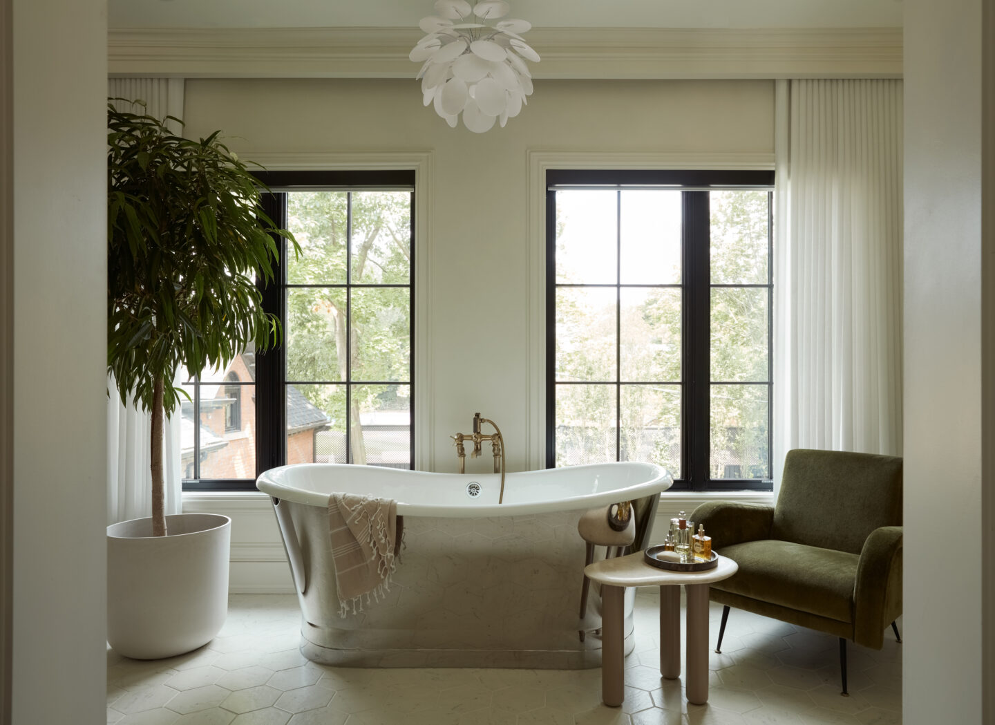 A freestanding tub with a plant to the left, a comfy chair to the right, and a tray of oils and bottles.