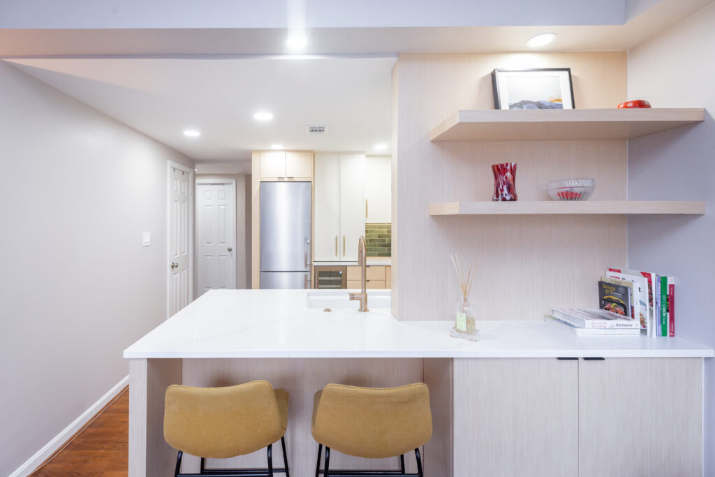 Kitchen peninsula with white quartz countertop, two mustard stools, light wood cabinetry, and floating shelves styled with décor and cookbooks.