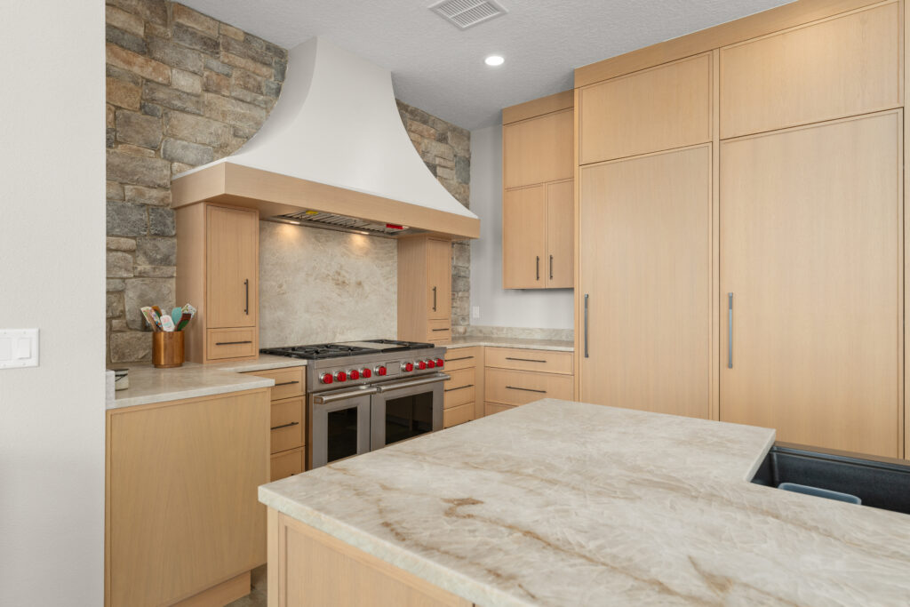 Custom kitchen remodel featuring light wood cabinetry, a stone accent wall with a sculpted white range hood, a professional-grade gas range with red knobs, and a large stone island with waterfall-style edges.