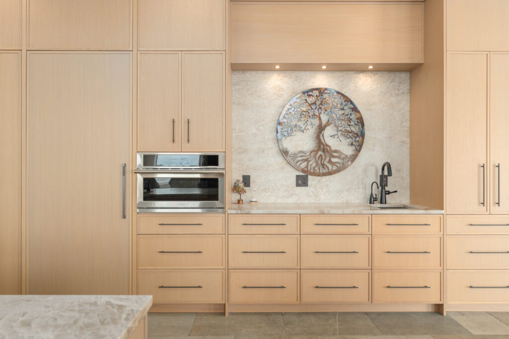 Custom kitchen remodel featuring light wood cabinetry, a stone accent wall with a sculpted white range hood, a professional-grade gas range with red knobs, and a large stone island with waterfall-style edges.