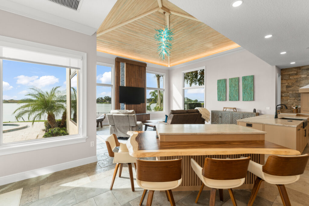 Open-concept kitchen and living area remodel featuring a large stone island with integrated sink, light wood cabinetry, a stone-accented range wall with sculpted hood, vaulted wood ceiling with decorative lighting, and expansive windows overlooking a waterfront view.