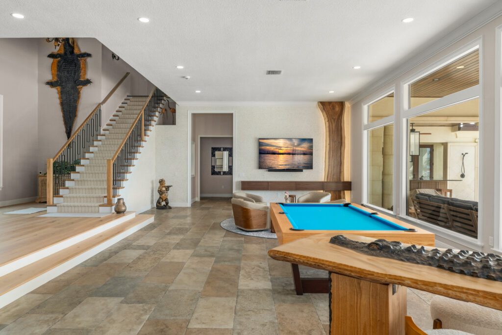 Spacious open living area remodel featuring a custom staircase with wood treads and black metal railing, a seating area with floating wood media console and mounted TV, a live-edge wood accent detail, large windows opening to an outdoor space, and a custom pool table anchoring the room.