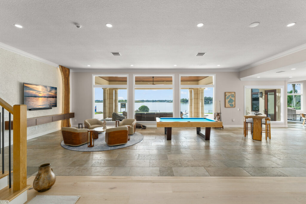 Spacious open living area remodel featuring a custom staircase with wood treads and black metal railing, a seating area with floating wood media console and mounted TV, a live-edge wood accent detail, large windows opening to an outdoor space, and a custom pool table anchoring the room.