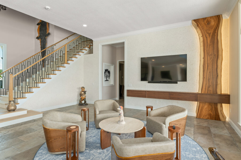 Spacious open living area remodel featuring a custom staircase with wood treads and black metal railing, a seating area with floating wood media console and mounted TV, a live-edge wood accent detail, large windows opening to an outdoor space, and a custom pool table anchoring the room.