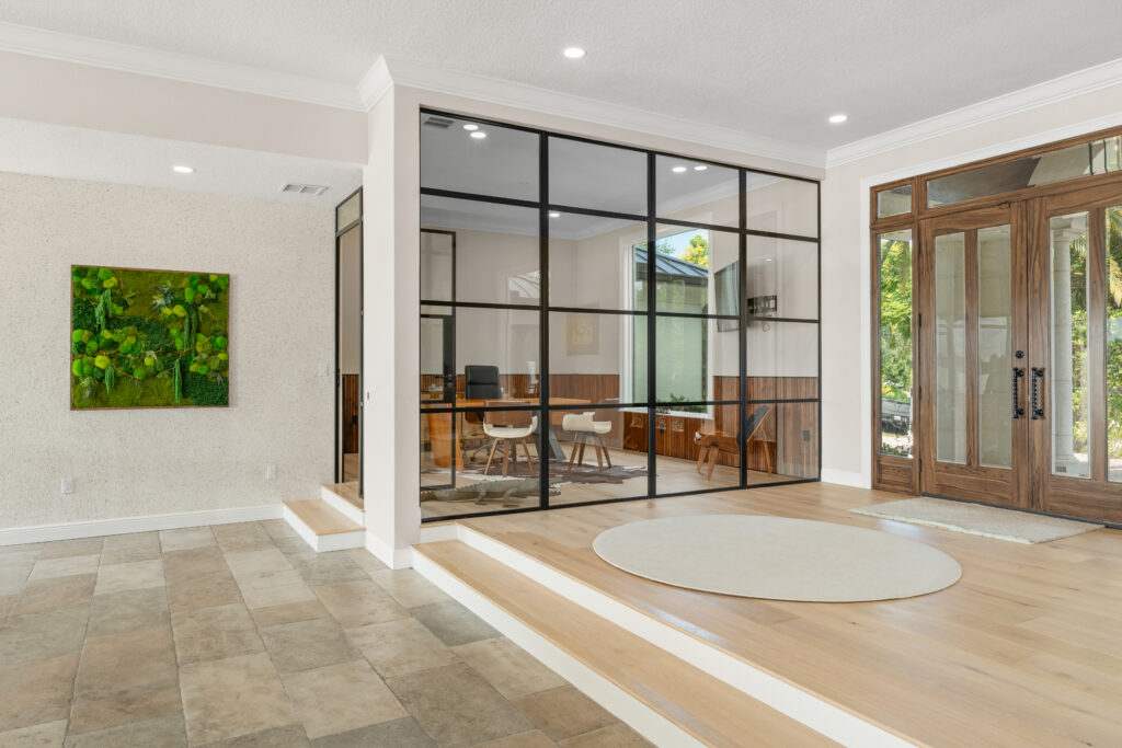 Modern home remodel featuring a glass-walled office with black metal framing, wood flooring with illuminated step detail, a textured accent wall with artwork, and large wood-framed entry doors bringing in natural light.