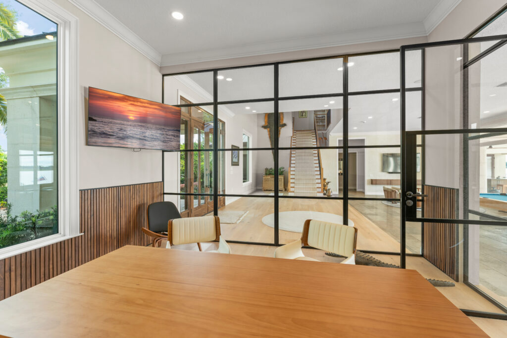 Modern home remodel featuring a glass-walled office with black metal framing, wood flooring with illuminated step detail, a textured accent wall with artwork, and large wood-framed entry doors bringing in natural light.