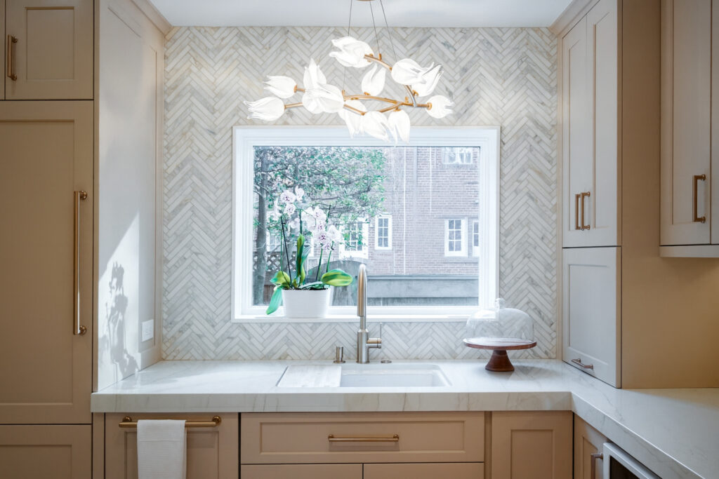 Kitchen sink framed by a large window with marble-look herringbone backsplash, quartz countertops, and contemporary pendant lighting.