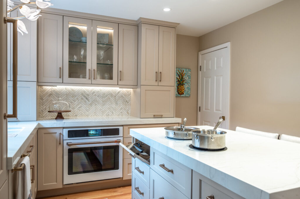 Modern kitchen with built-in wall oven, glass-front upper cabinets, herringbone tile backsplash, and quartz island countertop.