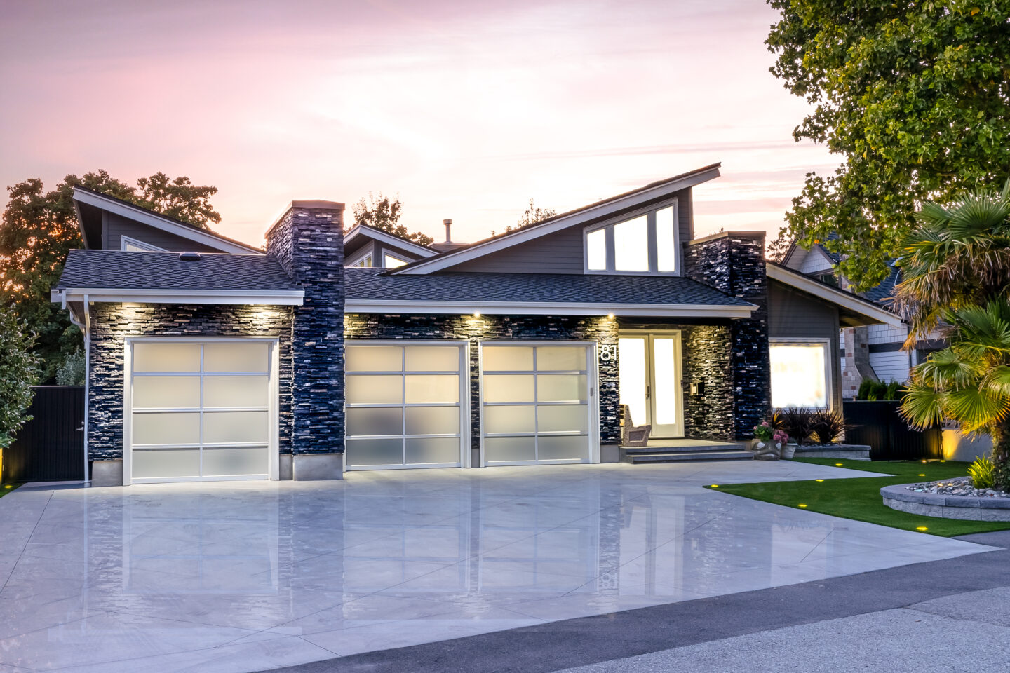 Modern custom home exterior with dark ledgestone cladding, frosted glass garage doors, angled rooflines, and a polished concrete driveway at dusk.