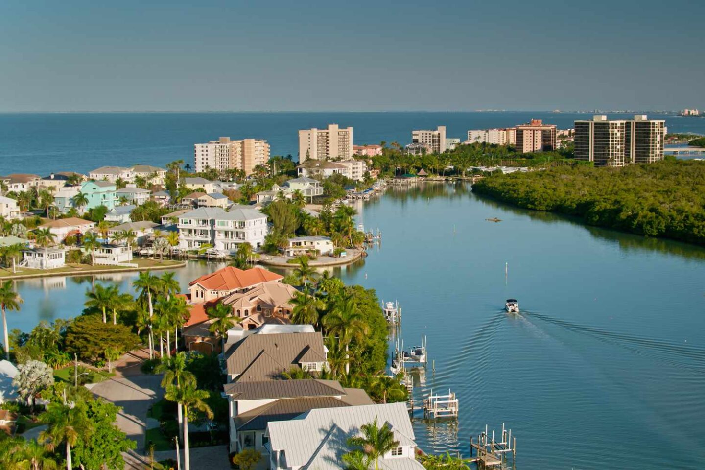 Aerial view of a coastal Florida neighborhood with waterfront homes, palm trees, private docks, and a calm bay leading to the Gulf of Mexico.