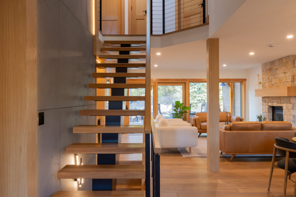 Open-riser wood staircase in a Bow Valley home renovation connecting a mountain-modern living space with a stone fireplace and natural light, designed to improve circulation, visual openness, and long-term home value in Canmore.