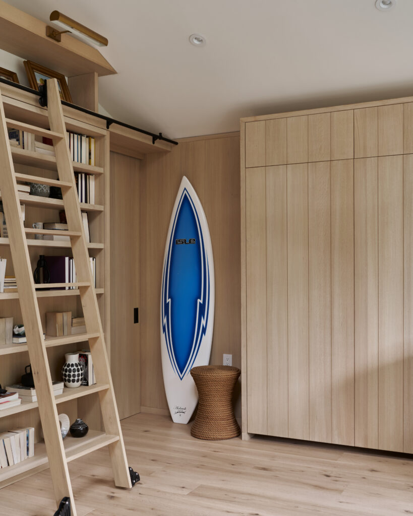 Wooden paneling room with a bookcase and sliding ladder to the left and a surfboard placed upright in the center of the room.