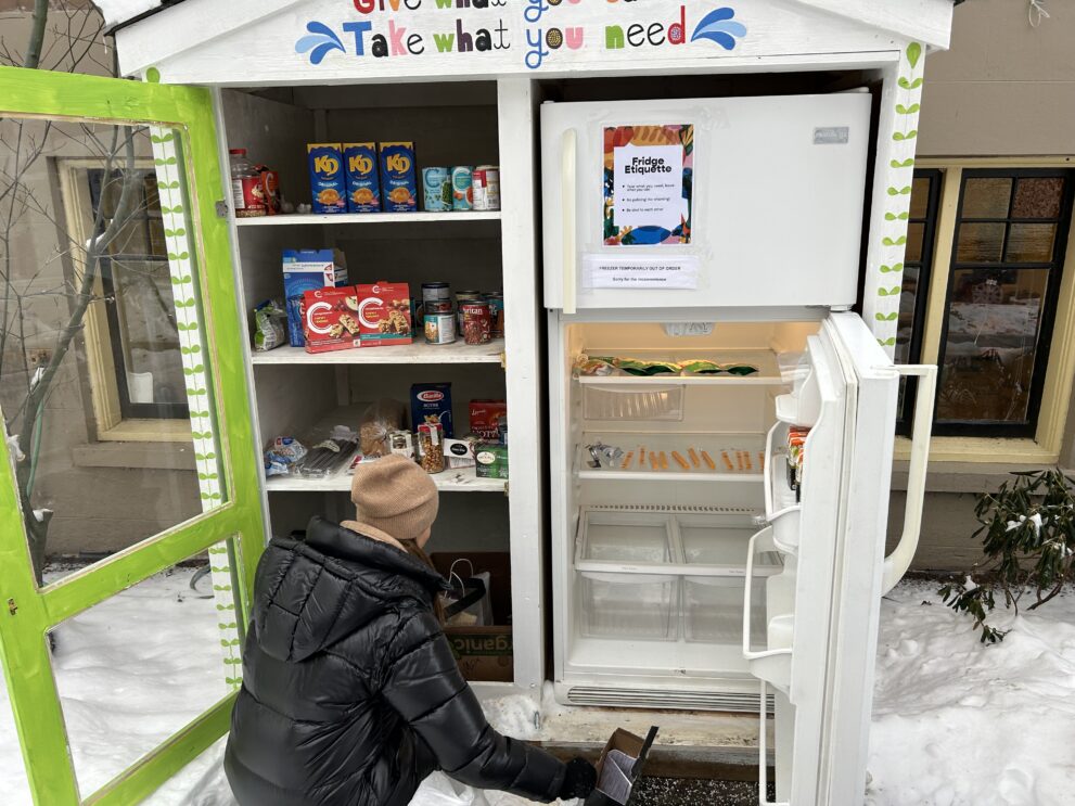 A donor in a winter coat and knit hat crouches to reach the bottom shelf of the food pantry, which is filled with cans and boxes of non-perishable foods.