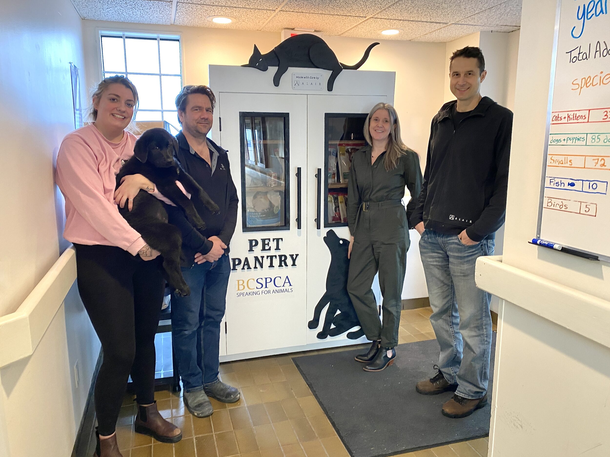 Four people (including a woman holding a black cat) stand beside a white food pantry labelled 