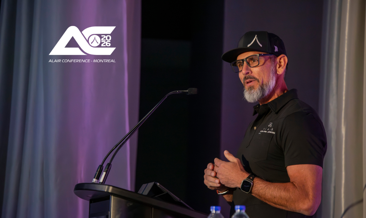Kevin Scott, wearing Alair-branded hat and shirt, speaks at a podium lectern. The top left logo reads 