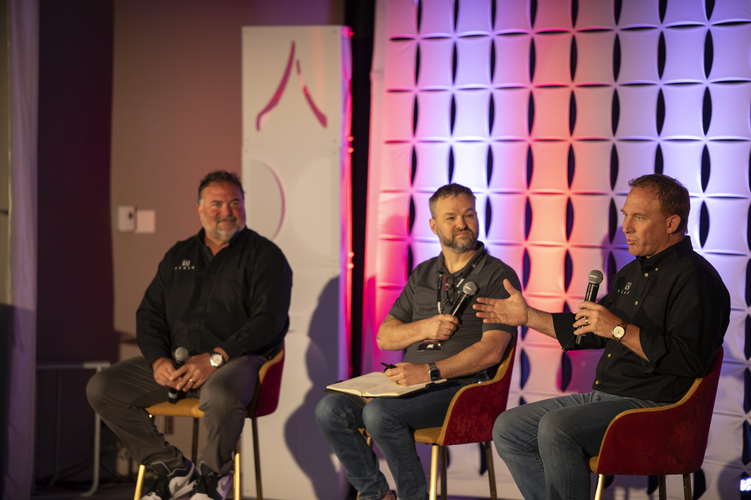 Three male speakers sit on a panel at the 2026 Alair Conference sitting under the Alair logo.