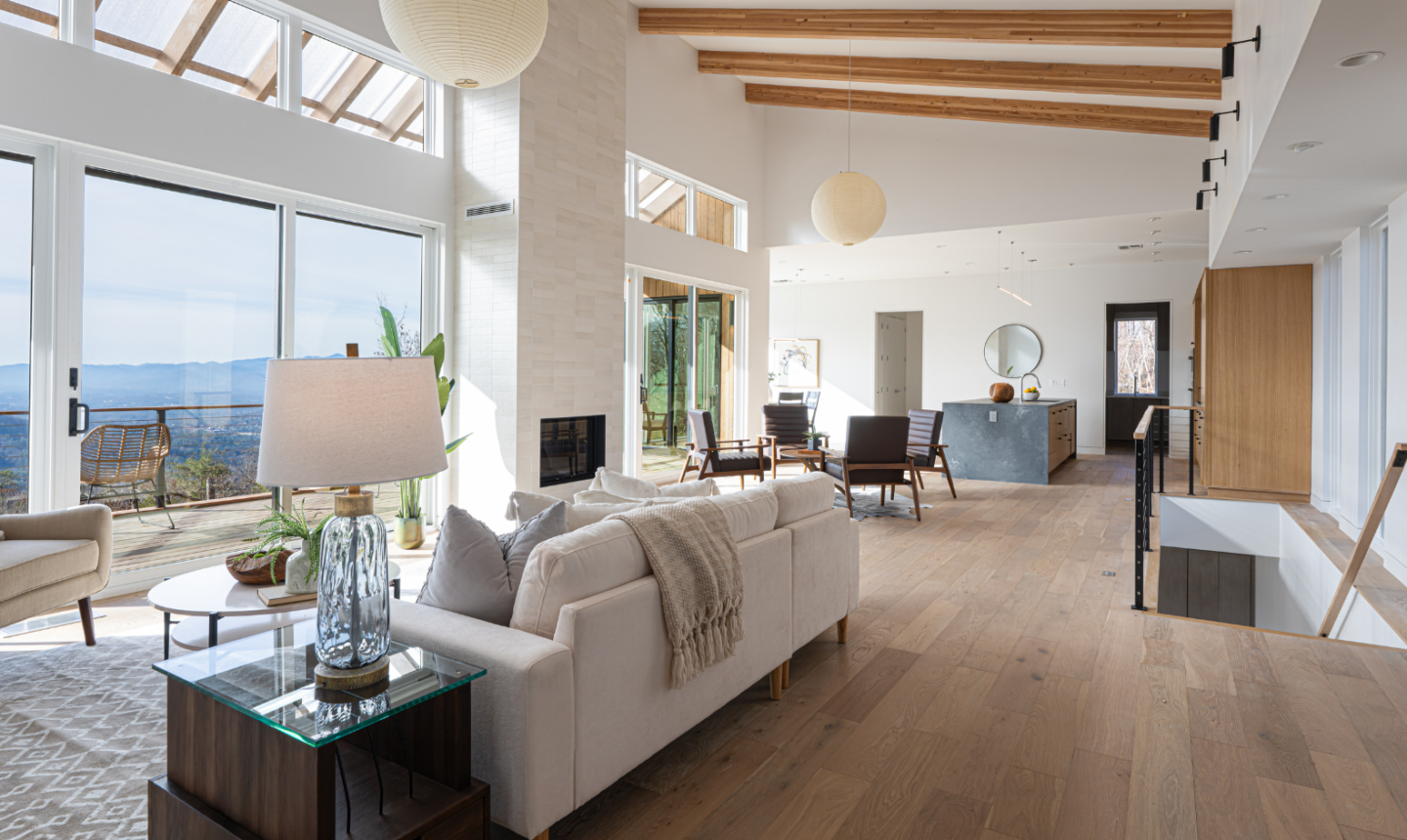 Modern open-concept living space with large floor-to-ceiling windows and mountain views, featuring natural wood beams, light wood flooring, neutral furnishings, a fireplace, and a kitchen island in a bright, sunlit interior that emphasizes connection to the outdoors.