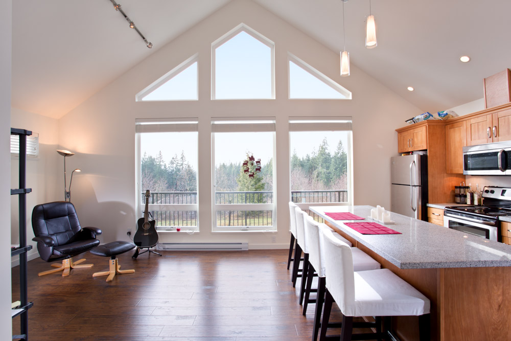 Interior of Vancouver coach home open-concept living room, showing floor-to-ceiling windows, a leath chear, a guitar, and a kitchenette.