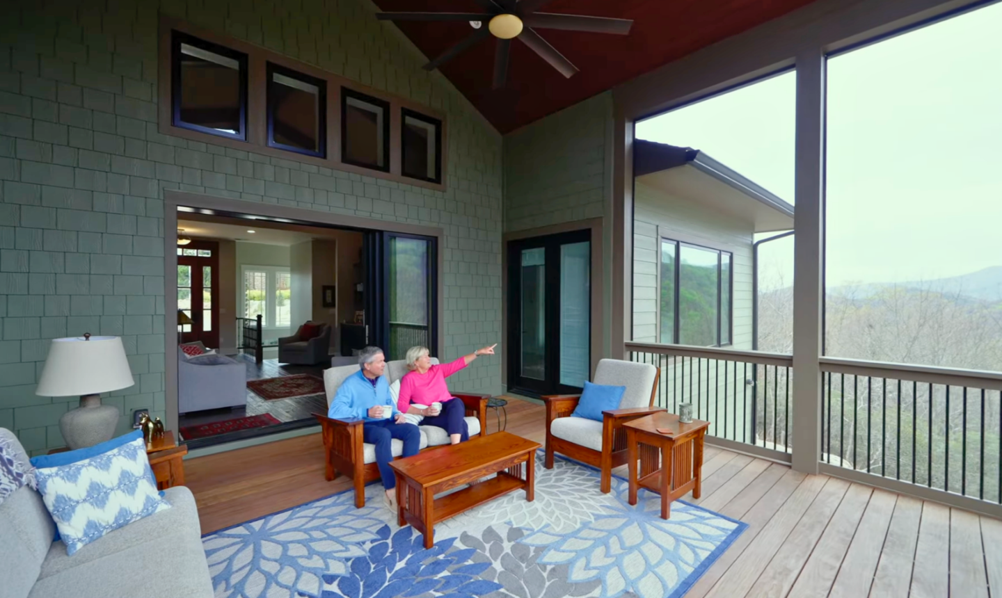 Older couple sitting together on a wooden sunroom on the second story of the house. The porch is spacious, with large windows offering a scenic view of distant hills and trees. The couple is seated on a wooden bench with blue cushions, and the woman is pointing out toward the view while the man looks in the same direction, both holding mugs. The porch is furnished with comfortable seating, a coffee table, a side table with a lamp, and a patterned blue and white rug. The atmosphere is cozy and inviting, suggesting a peaceful and accessible home environment ideal for aging in place.