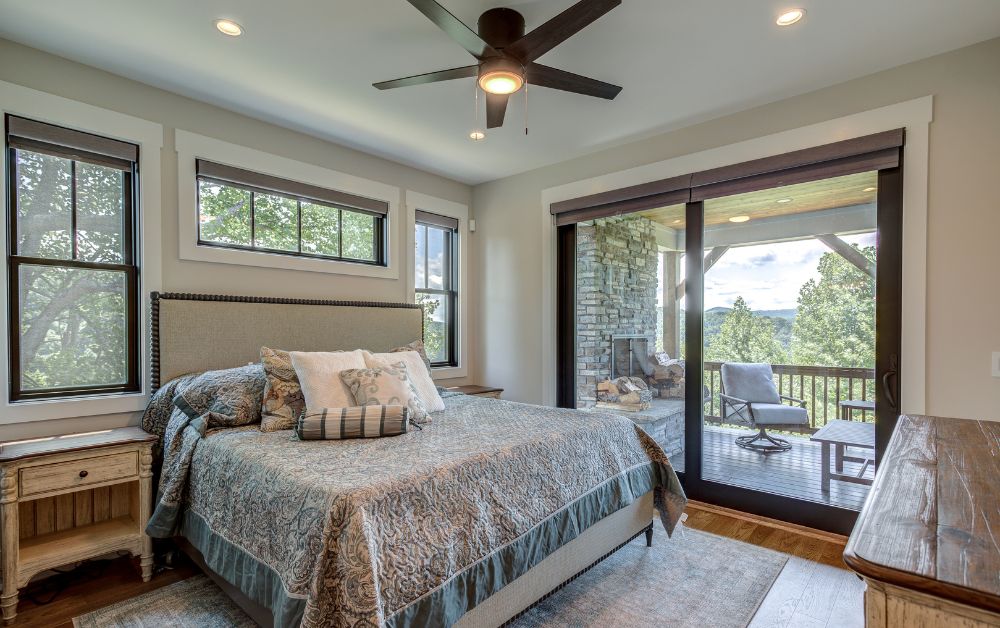 Primary bedroom in a custom High Country home featuring large windows, a king bed, hardwood floors, and sliding glass doors that open to a covered mountain-view porch with a stone fireplace.