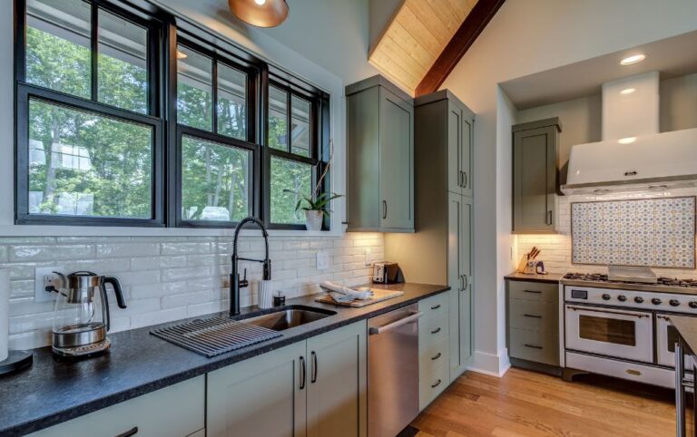 Bright custom kitchen with sage green cabinets, black windows, quartz countertops, wood ceiling details, and a designer tile backsplash in a High Country mountain home.