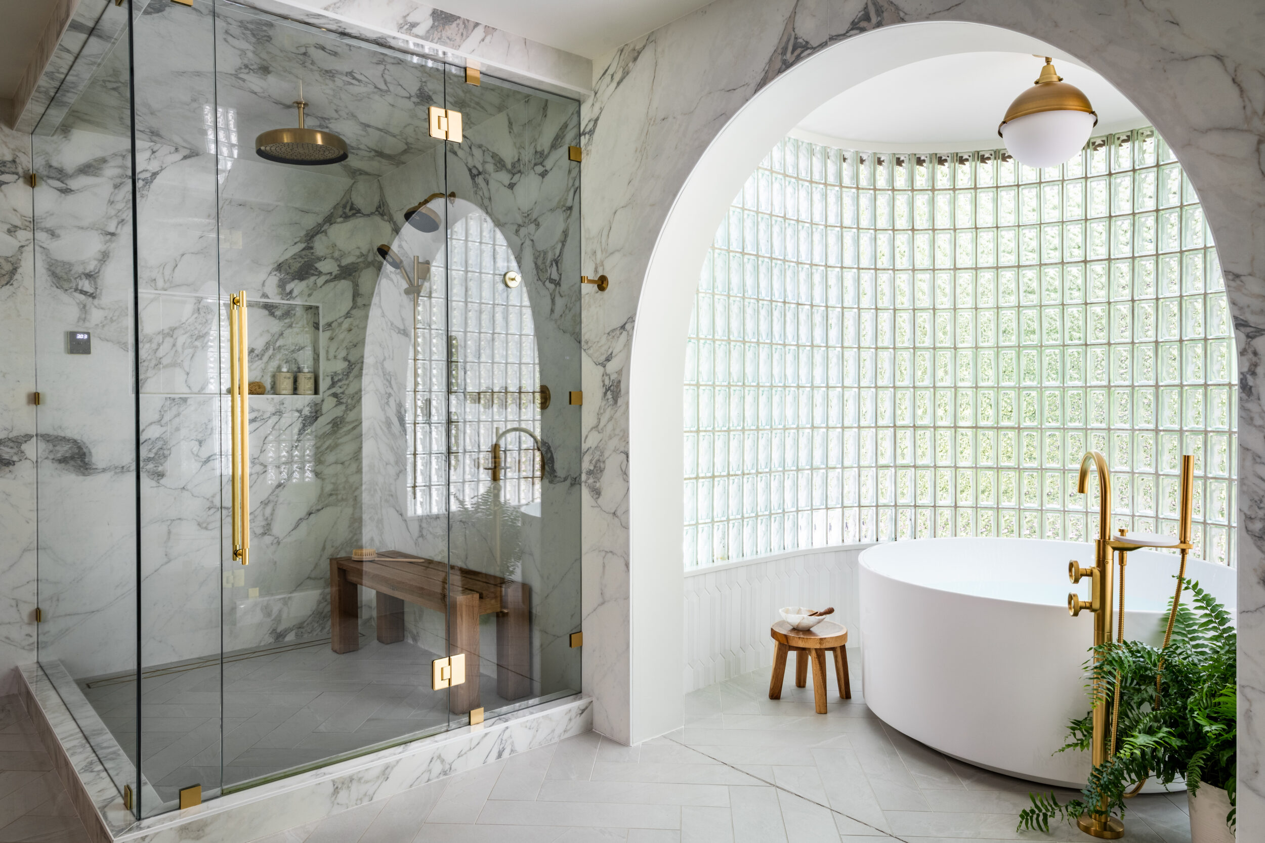 Luxury primary bathroom featuring a curved glass block wall, freestanding soaking tub, marble-clad steam shower, brass fixtures, and a dramatic arched opening. Provide your feedback on BizChat