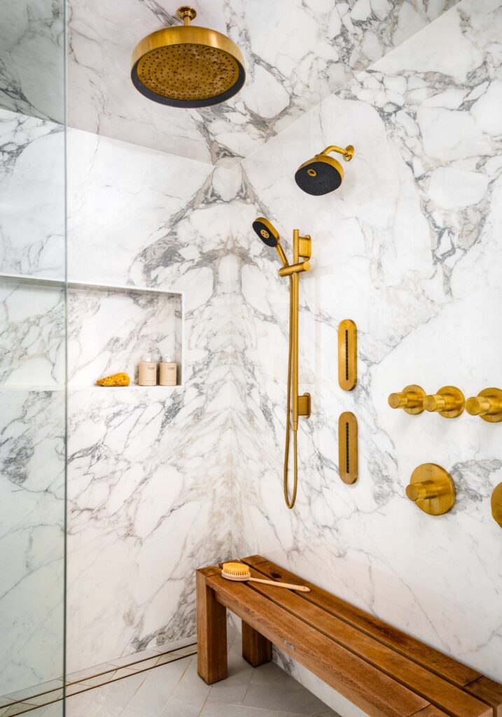 Marble-clad steam shower with brass fixtures, dual showerheads, built-in bench seating, and dramatic bookmatched stone walls in a luxury primary bathroom.