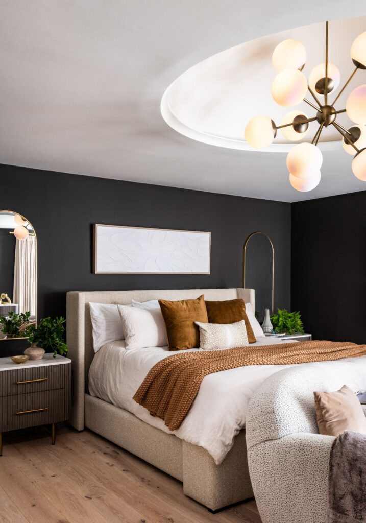 Contemporary primary bedroom with a dark accent wall, upholstered bed, warm wood flooring, brass lighting, and a statement chandelier set within a recessed ceiling detail.