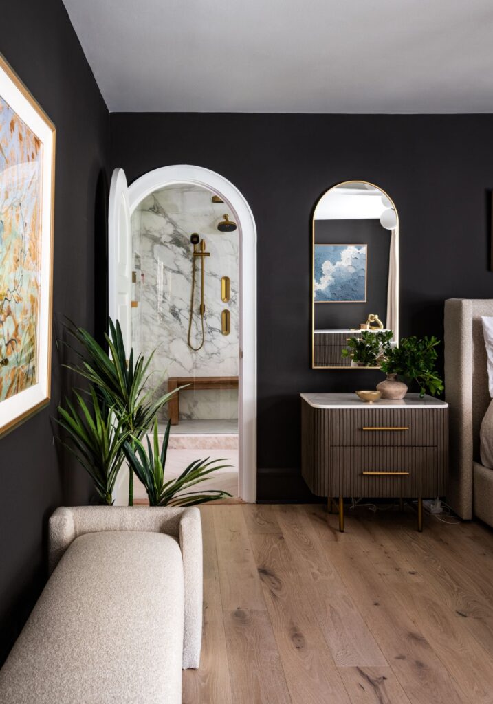 Primary bedroom sitting area with dark walls, warm wood flooring, arched doorway, and views into a marble-clad spa bathroom with brass fixtures and a steam shower.