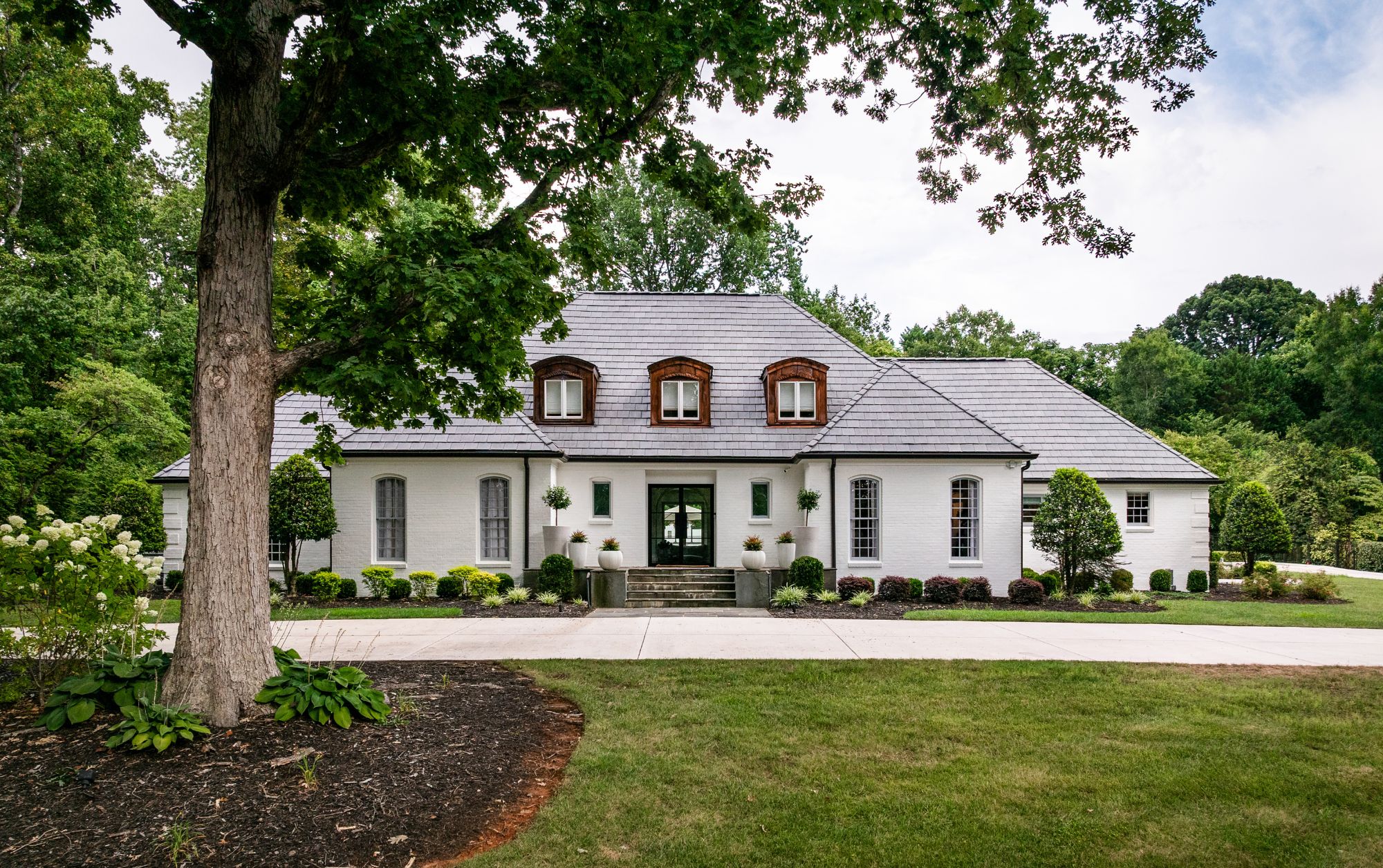 Front exterior of a luxury renovated home featuring painted brick, dormer windows, a slate-style roof, manicured landscaping, and mature trees on a private lot.