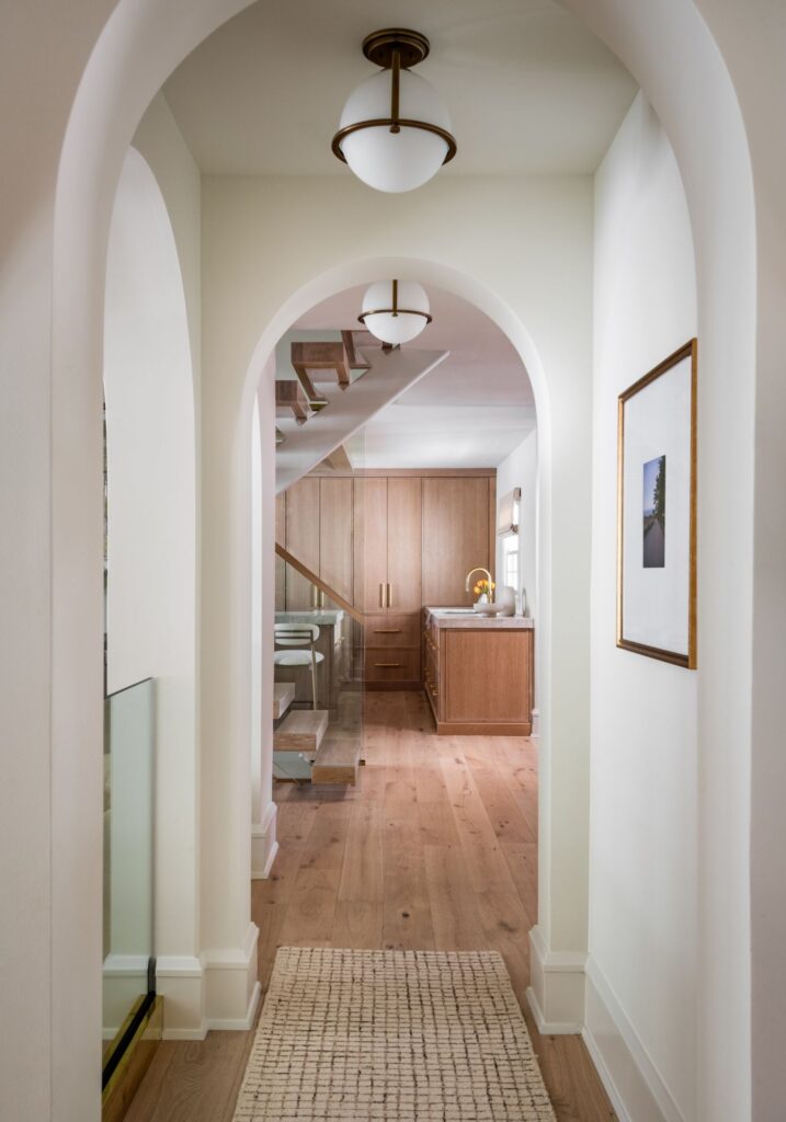 Arched hallway with warm wood flooring, globe pendant lighting, custom millwork, and views of a glass-enclosed staircase beyond.