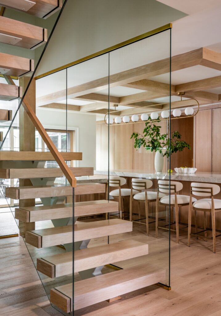 Glass-enclosed staircase with floating wood treads overlooking a custom kitchen featuring warm wood cabinetry, a waterfall island, and white oak ceiling beams.