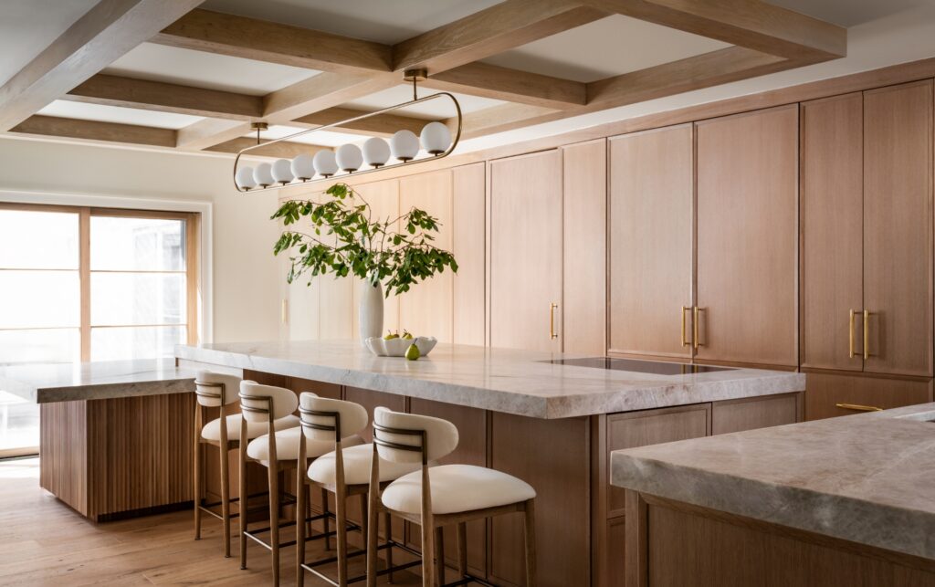 Contemporary kitchen with a waterfall-edge stone island, custom wood cabinetry, brass hardware, coffered wood ceiling, and upholstered counter stools beneath a modern globe chandelier.