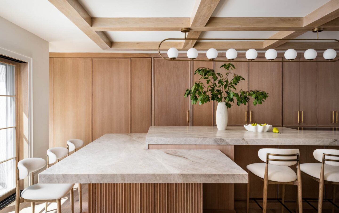 Custom kitchen with dual waterfall-edge stone islands, warm wood cabinetry, brass hardware, coffered wood ceiling beams, and modern upholstered seating in a light-filled open-concept layout.