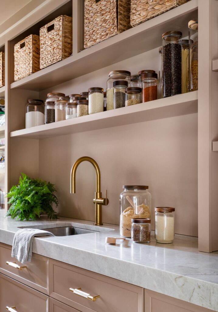 Custom scullery with open shelving, woven storage baskets, labeled glass pantry jars, brass faucet, and stone countertops designed for organized kitchen storage and prep.