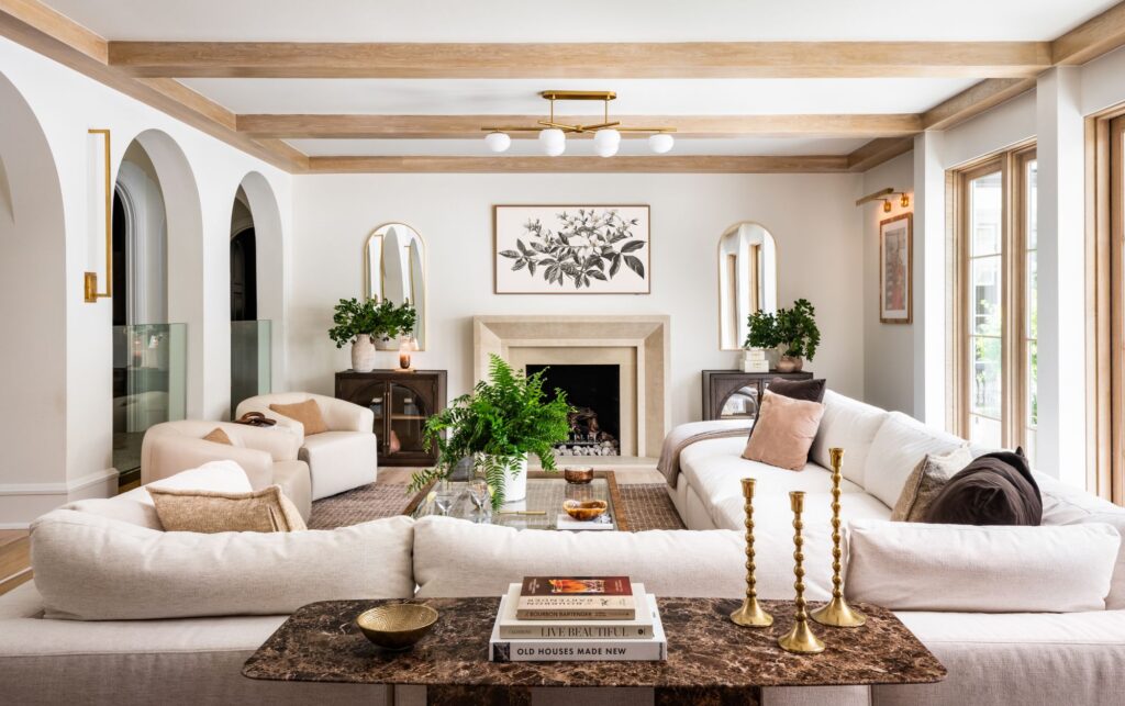 Bright living room with a stone fireplace, arched architectural details, white oak ceiling beams, glass railings, and oversized windows that fill the space with natural light.