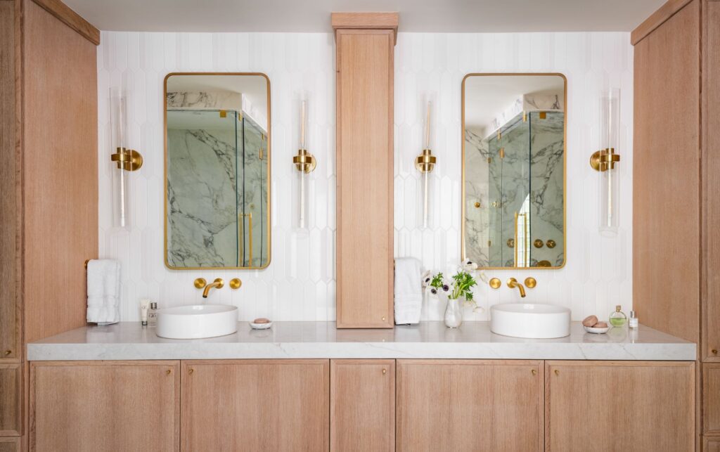 Double vanity with warm wood cabinetry, vessel sinks, brass fixtures, and arched mirrors, reflecting a marble-clad steam shower in a spa-inspired primary bathroom.