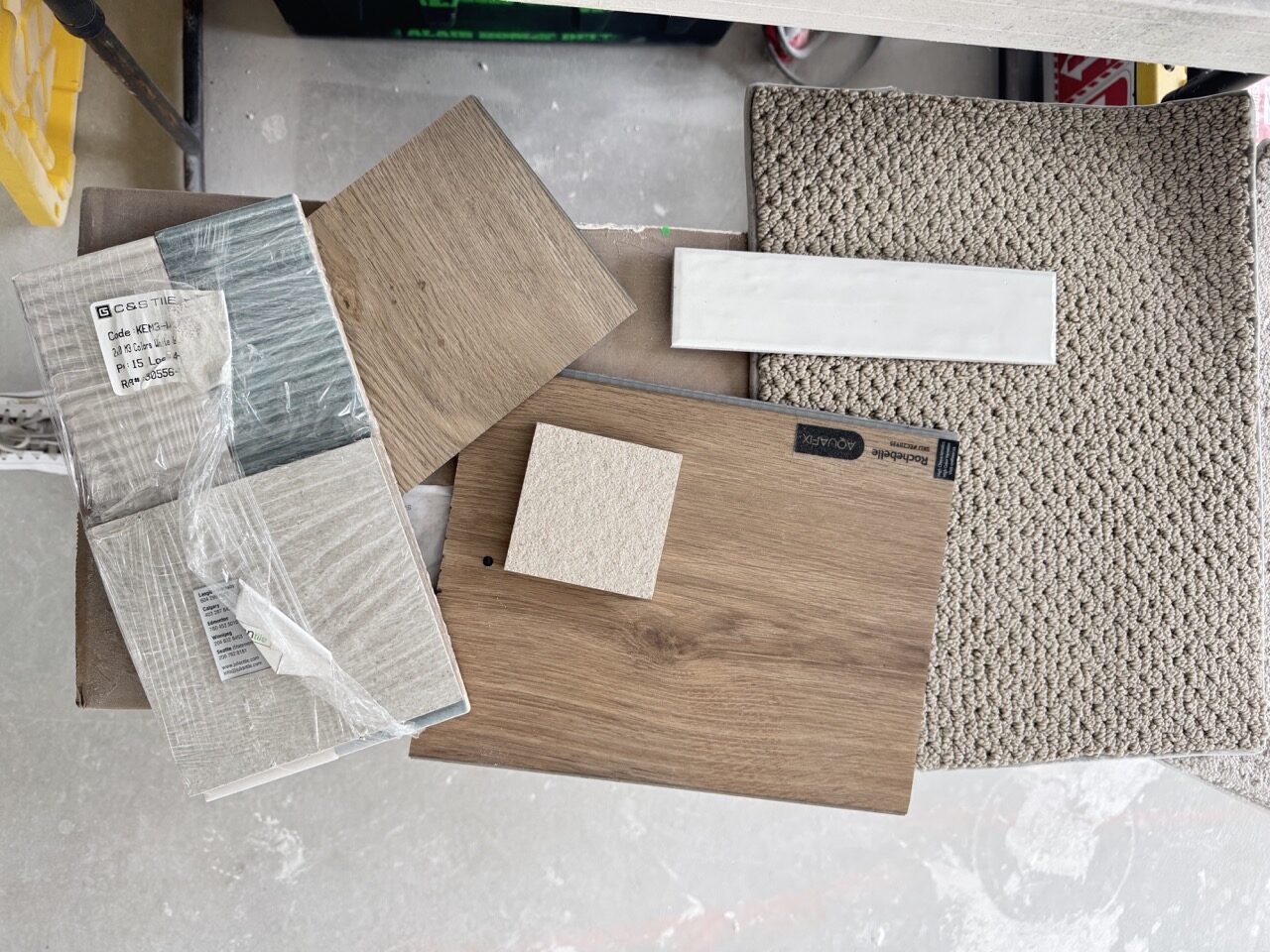 Flooring and finish samples arranged on site, including textured loop carpet, white subway tile, wide-plank oak vinyl flooring, and stone-look porcelain tile